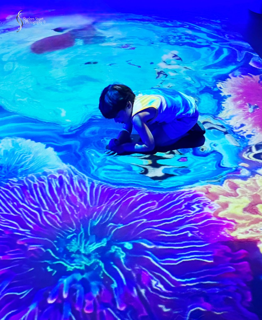 A child crouching on a colorful, animated floor display featuring swirling patterns in blue, purple, and pink hues.