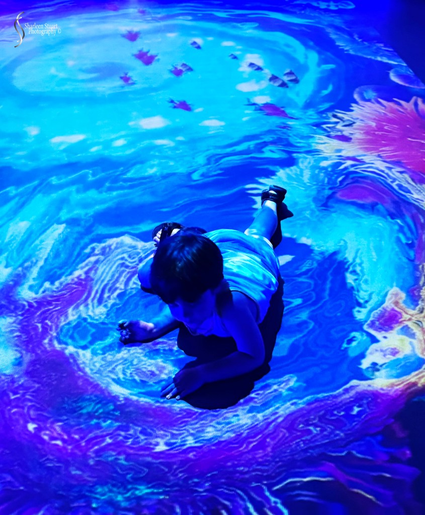 A child lying on a colorful, projected floor depicting swirling blue and purple patterns, creating an immersive artistic environment.